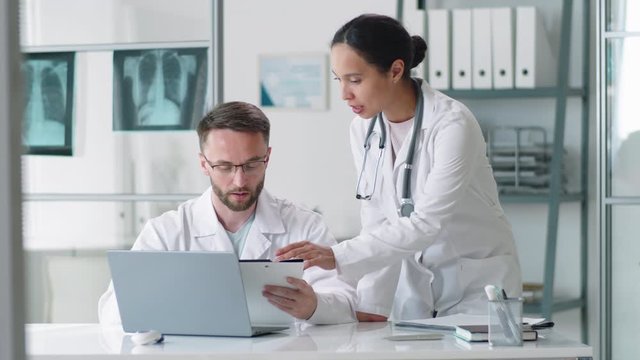 Young brunette female doctor walking to male colleague, leaning on desk, giving him clipboard and discussing document on it while working together in medical office