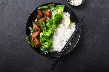 Vegan teryaki  tempeh or tempe buddha bowl  with rice, steamed broccoli, spinach  and lime on black  background. Healthy food