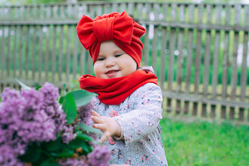 little girl in a red hat with a bow is in the yard near the purple lilac