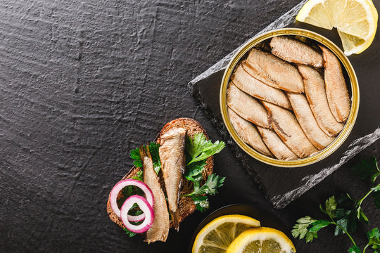 Sprats in tin can and sandwich with sardines, sprats fish with parsley, lemon, spices on rye bread over slate dark background. Mediterranean food, appetizer, seafood, top view
