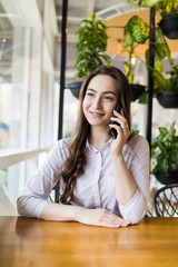 Young happy woman talking on mobile phone with friend while sitting in modern coffee shop