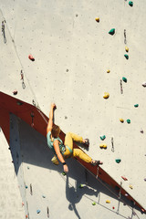 woman climber in bright yellow pants climbing on artificial rock wall in climbing gym, resting and...