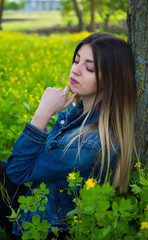 A girl in denim clothes with beautiful hair sits leaning on a tree with a dreamy look on her face. Warm spring day