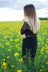 Naklejka na ściany i meble Girl in dark clothes with beautiful hair standing in a field of yellow rape. Warm spring day
