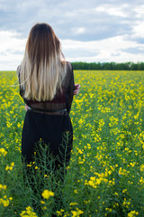 Fototapeta premium Girl in dark clothes with beautiful hair standing in a field of yellow rape. Warm spring day
