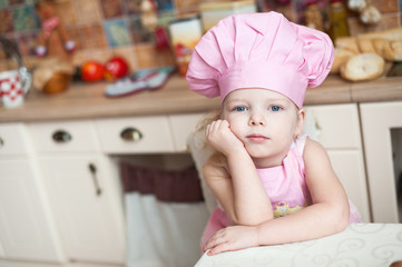 little girl in the kitchen in a pink chef's hat and apron sits at the table, pensive, dreamy.