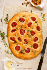 Italian traditional focaccia bread baking with with cherry tomatoes, parmesan and rosemary on light brown background. Top view.