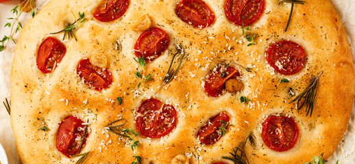 Italian traditional focaccia bread baking with with cherry tomatoes, parmesan and rosemary on light brown background. Top view.