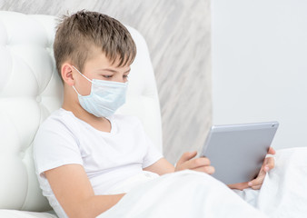 Ill boy wearing medical protective mask sits on the bed at home and uses tablet computer. Quarantine and coronavirus epidemic concept. Empty space for text