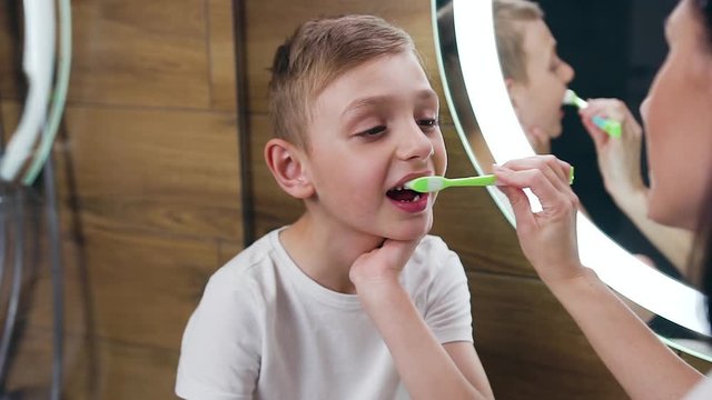 Likable high-spirited 10-aged blond boy sitting near the bathroom mirror with open mouth as her happy pretty mother brushing his teeth and kissing him to nose