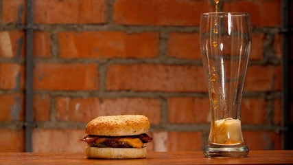 Juicy tasty burger lies on wooden table. Beer is poured into a glass. Jet of craft dark beer breaks into drops on the walls of a glass with bubbles and foam rise. Unhealthy foods in self-isolation 