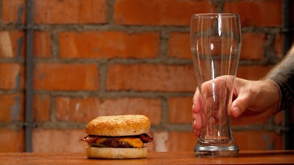 Man's hand holds a empty glass. Tasty beef burger with cheese and golden bun on wooden table on brick wall background. Unhealthy quarantine foods. 