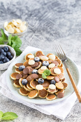 Tiny pancakes for breakfast. Cereal pancakes with blueberries and chocolate drops in plate on grey background. Trendy food. Copy space for text or design