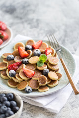 Tiny pancakes for breakfast. Cereal pancakes with blueberries, strawberries in plate on grey background. Trendy food. Selective focus