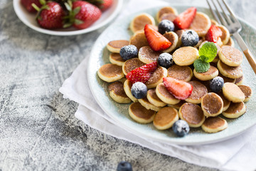 Tiny pancakes for breakfast. Cereal pancakes with blueberries, strawberries in plate on grey background. Trendy food. Selective focus