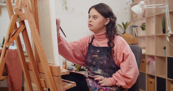 Young talanted teenager wearing artists apron painting while sitting in front of molbert at her room. Pretty girl with genetic disorder holding palette on her knees creating picture.
