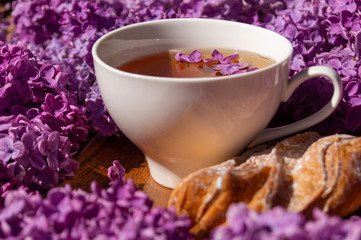 white porcelain cup with tea and a crispy bun among flowering branches of lilac