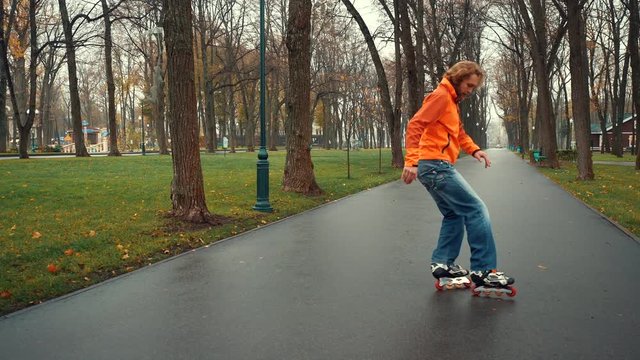 Technical and experienced skater making complex turns and feints on skates in an alley of city park among trees in wet weather. Active leisure sporty bearded man on vacation or weekend.