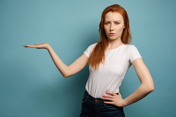 The red-haired girl stands on a blue background in a white T-shirt, points with the palm to the side and frowns at the frame