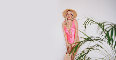 Young sexy smiling happy blonde in a pink swimsuit, a straw hat, sunglasses, excited to present product. Woman on a white background with palm green leaves. Summer sales concept copy space mock up