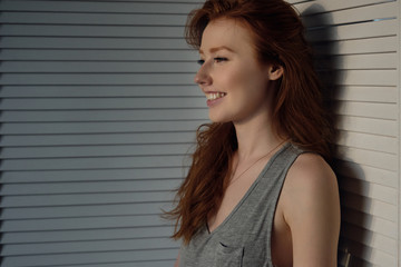 Redhead girl with freckles stands in front of a white wooden screen in profile and smiles.