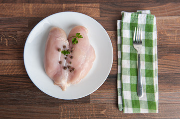 Fresh raw chicken fillet and parsley on a white plate on a wooden background, with a fork and towel, top view. 
Chicken fillet with seasoning ready to cook. proper nutrition.
