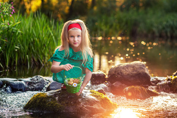 A girl in a green dress sits on a rock in the river. A blond kid with a red headband and red shoes is holding a basket. the river in the sun.