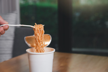 Hand of a man holding a plastic fork with cooked instant noodles. A cup of instant noodle with fork...