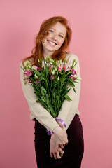 A cute red-haired girl stands on a pink background, hugging a bouquet of flowers smiling widely.