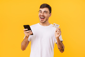 Photo of delighted young man holding credit card and cellphone