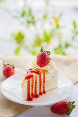 Close up strawberry crepe cake sliced on a white plate decorated with strawberry fruits on a wite table with strawberry and green nature background.