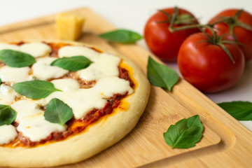 Homemade classic Italian Napoli pizza with tomato sauce, mozzarella cheese and basil leaves: pizza Napoletana. It is on a wooden cutting board surrounded by tomatoes, cheese and basil leaves