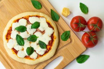 Homemade classic Italian Napoli pizza with tomato sauce, mozzarella cheese and basil leaves: pizza Napoletana. It is on a wooden cutting board surrounded by tomatoes, cheese and basil leaves