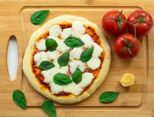 Homemade classic Italian Napoli pizza with tomato sauce, mozzarella cheese and basil leaves: pizza Napoletana. It is on a wooden cutting board surrounded by tomatoes, cheese and basil leaves