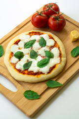 Homemade classic Italian Napoli pizza with tomato sauce, mozzarella cheese and basil leaves: pizza Napoletana. It is on a wooden cutting board surrounded by tomatoes, cheese and basil leaves