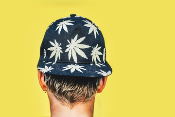 Young girl in sunglasses and cap with leaves of marijuana smokes on a yellow wall background. Hemp smoke puffs
