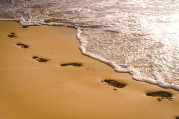 Sea waves hit the coast causing white bubbles and splashes of water. The orange color glow of the sunset creates warm tones. There are footprints on fine sandy beaches with glittering glitter.