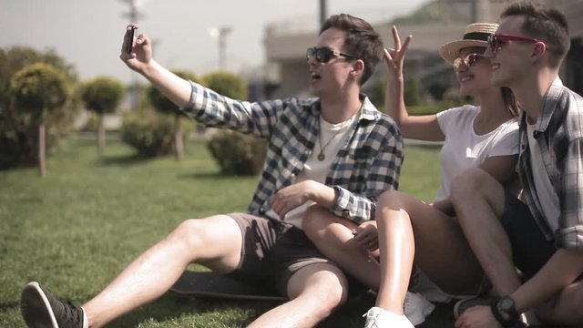 Three friends take a selfie while sitting on the grass in a modern city, traveling through the capitals of different countries. Travel concept. Friends concept. Prores 422. 