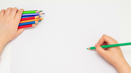 Top view of little hand of child drawing on blank paper by green pencil with bunch of colorful...