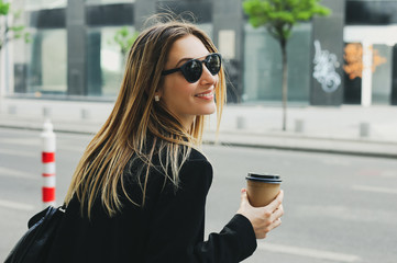 Portrait of young business woman walking on the street, in the morning with cup of coffee on her hand and wear sunglasses