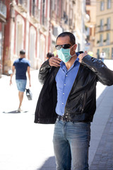 Uomo vestito casual e con mascherina fermo nel marciapiede di una città con paesaggio persone sullo sfondo