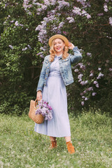 Cheerful young beautiful hipster woman in straw hat, denim jacket posing with purple lilac flowers...