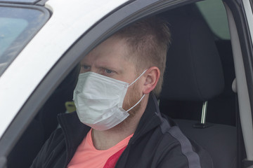 Fototapeta na wymiar Light man with blue eyes in a medical mask behind the wheel of a car