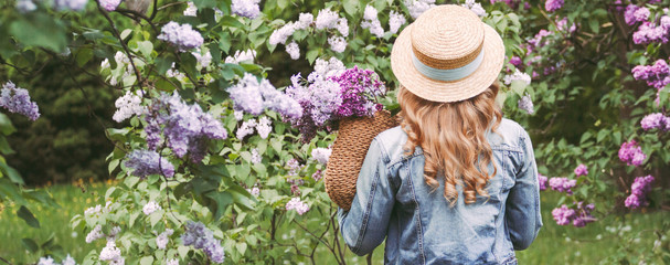 Panorama banner beautiful stylish young blonde woman in straw hat and denim jacket enjoy spring nature, relax in countryside lilac park. Happy attractive carefree hipster girl posing on blossom garden
