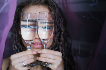 Young woman wearing dreadlocks holding and look throw vine glasses, big funny eyes optical distortion through water