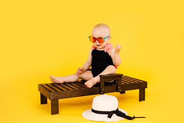 Funny little girl dressed in a black and pink swimsuit, big hat and sunglasses sitting on wooden deckchair isolated on yellow background. Summer holidays concept