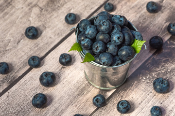 Close-up of a bucket with juicy fresh blueberries and small green leaves, berries scattered on the table.