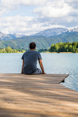 Fototapeta premium Mann sitzt auf einem Bootssteg mit einer schönen Alpenlandschaft und einem See