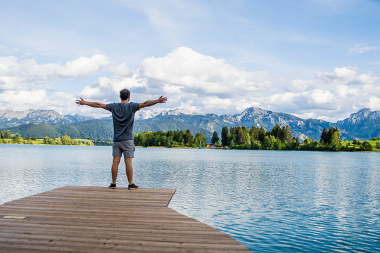 Mann steht auf einem Bootssteg mit ausgestreckten Armen und einer sch&ouml;nen Alpenlandschaft und einem See