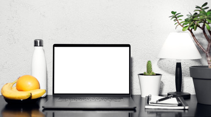 Workplace of freelancer, laptop with mockup on black desk beside lamp, steel thermo water bottle, pen on notebook, fruits and home flowers. Background of grey textured wall.